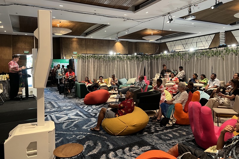 Audience members gather in a conference venue in Fiji to watch a video presentation hosted by Nicolas Gouletquer during a Pre-COP event. Attendees are seated on colourful beanbags, sofas and chairs facing a large presentation screen in a lounge-style setting.
