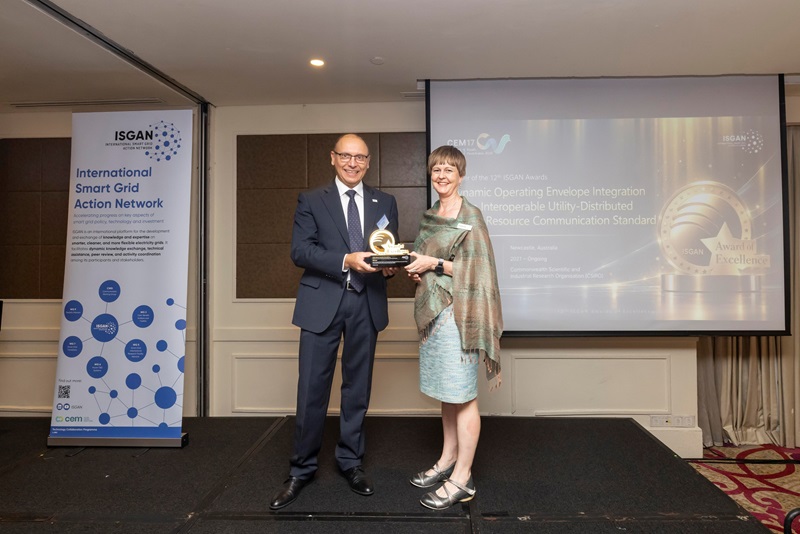 ISGAN Chair Luciano Martini presents the 2026 ISGAN Award of Excellence to Dr Gabrielle Kuiper, CSIRO's Australia-Southeast Asia DER lead. Both stand on stage holding the trophy, beside an International Smart Grid Action Network banner and in front of a presentation screen showing CSIRO's Dynamic Operating Envelope Integration project as the award winner.