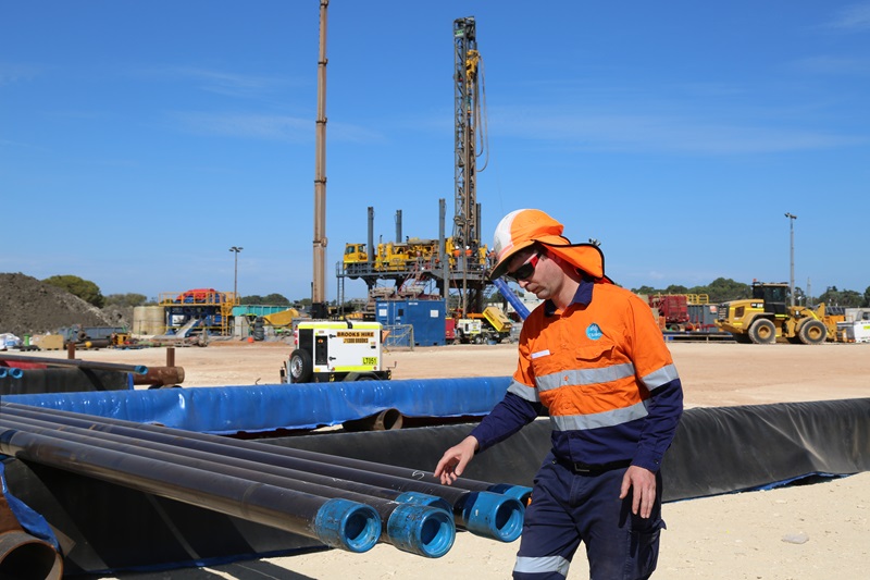 Person in high viz uniform and hard had on an industrial site, standing near industrial pipes with a rig-like structure in the background