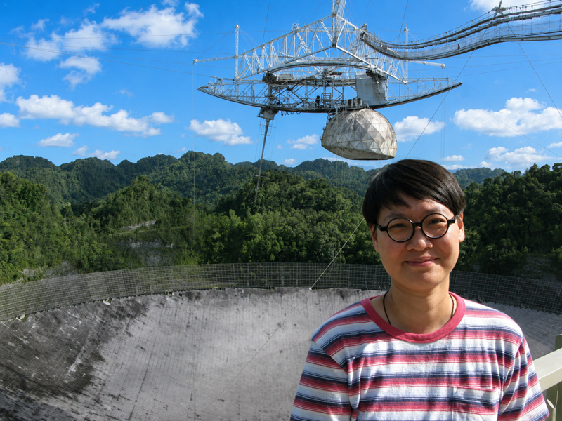 Woman stands in a striped shirt in front of satellite dishes as part of an observatory.