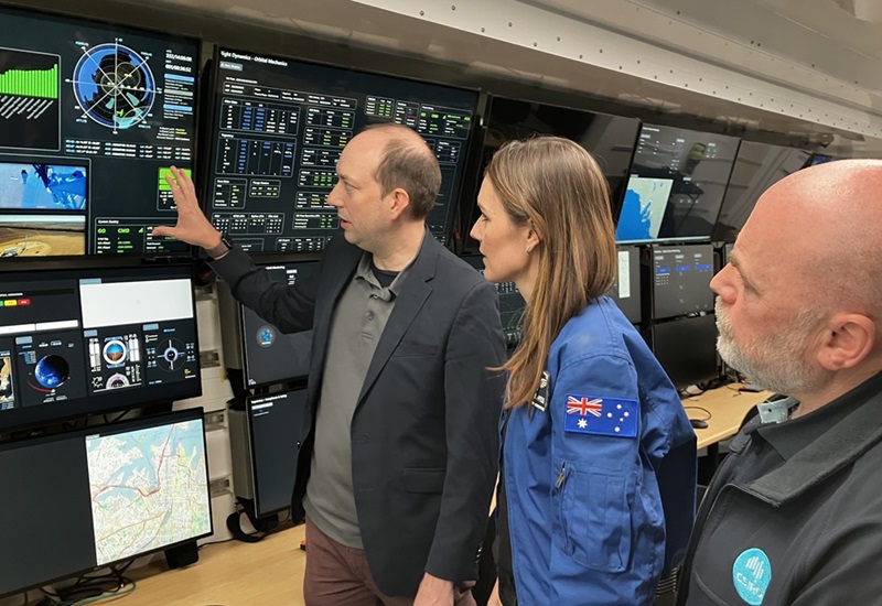 Katherine Bennell-Pegg and two men looking at wall of monitors.