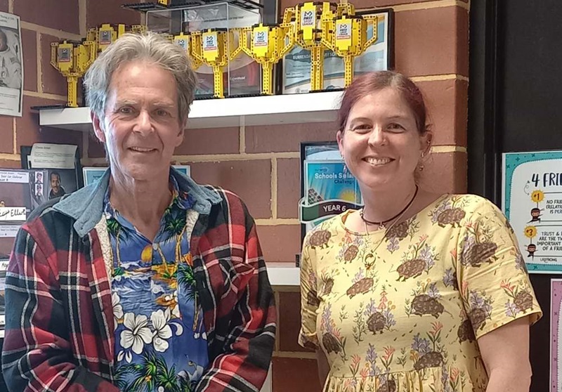 Two adults pose in front of shelves displaying STEM challenge trophies, awards and student achievement certificates in a classroom setting.