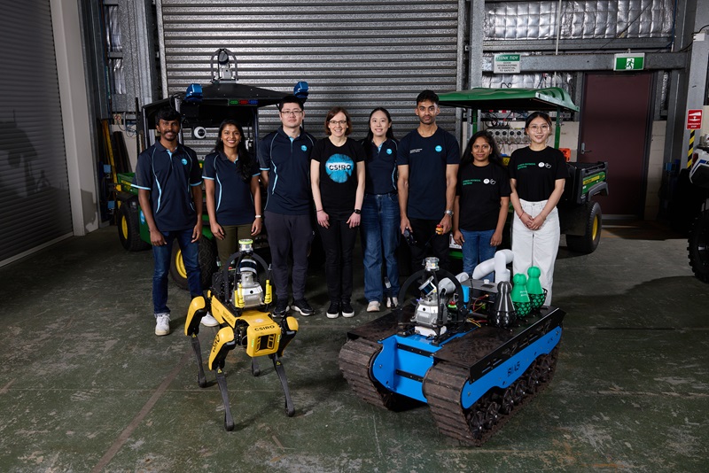 CSIRO Robotics researchers and engineers standing in a workshop with several autonomous robots, including a quadruped robot and a tracked ground robot.