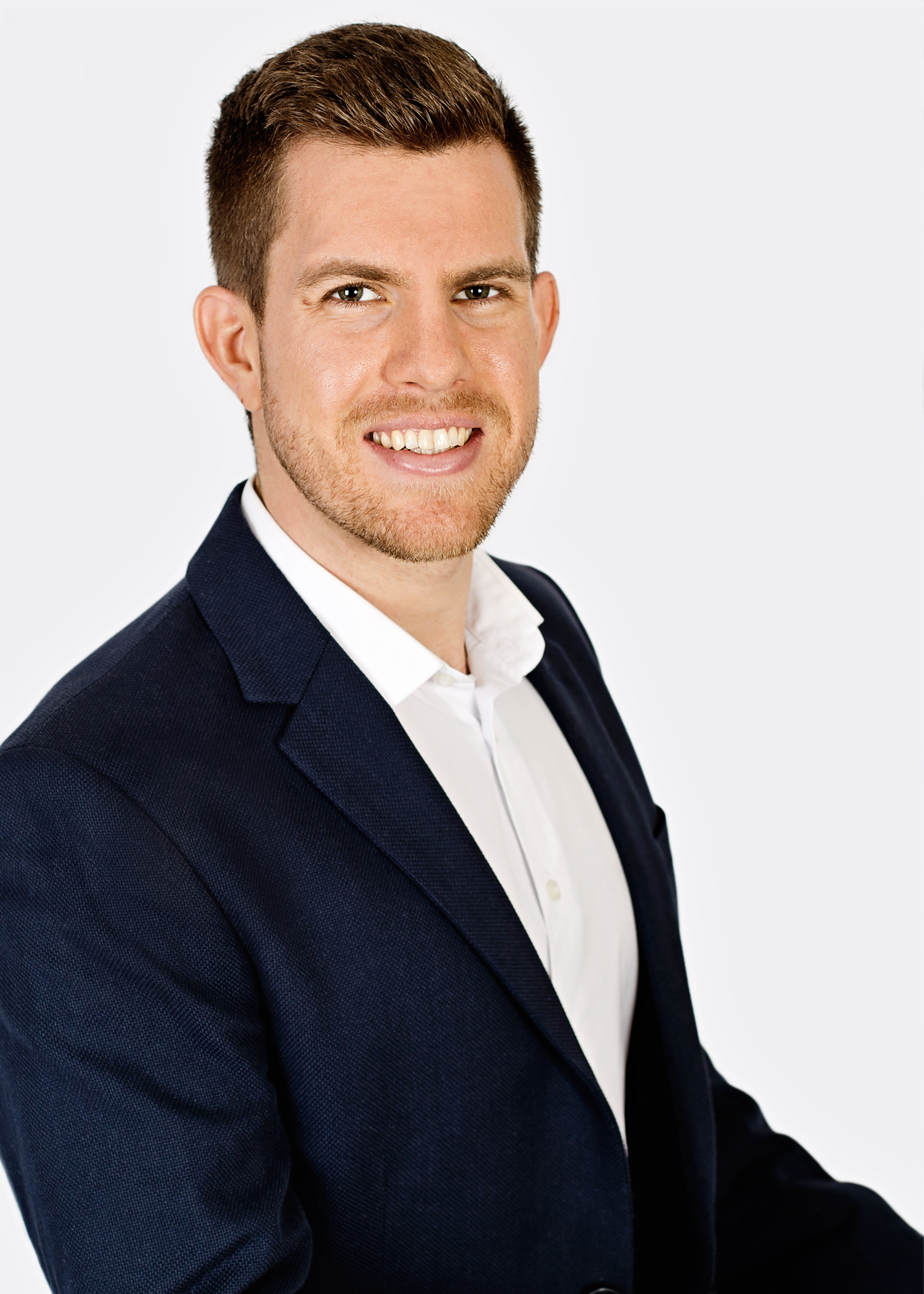 Male with short brown hair and slight facial hair. Corporate headshot, upper body. White shirt and dark blue blazer.