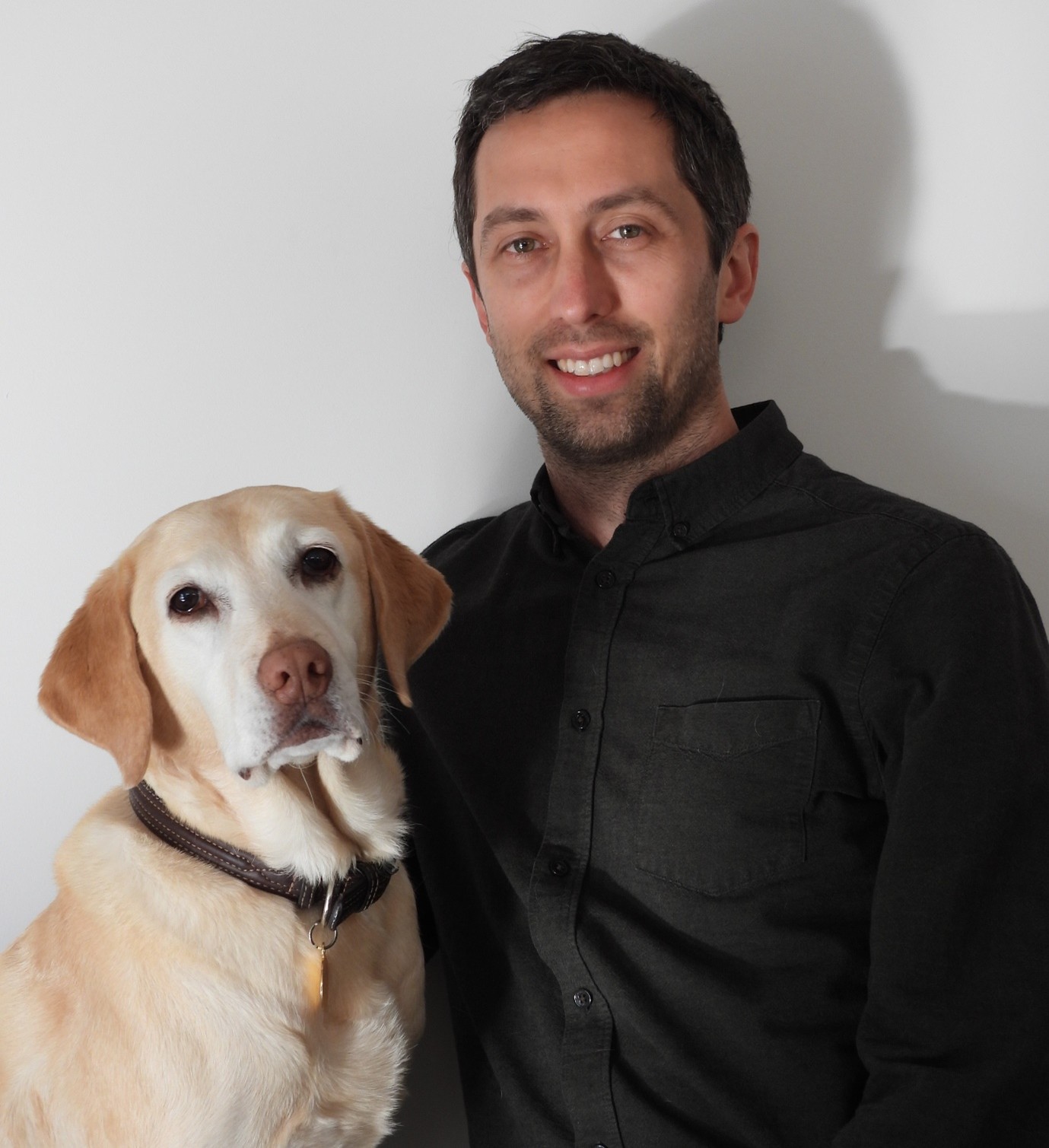 A yellow labrador retriever dog sitting next to a man with dark hair who is wearing a black shirt.
