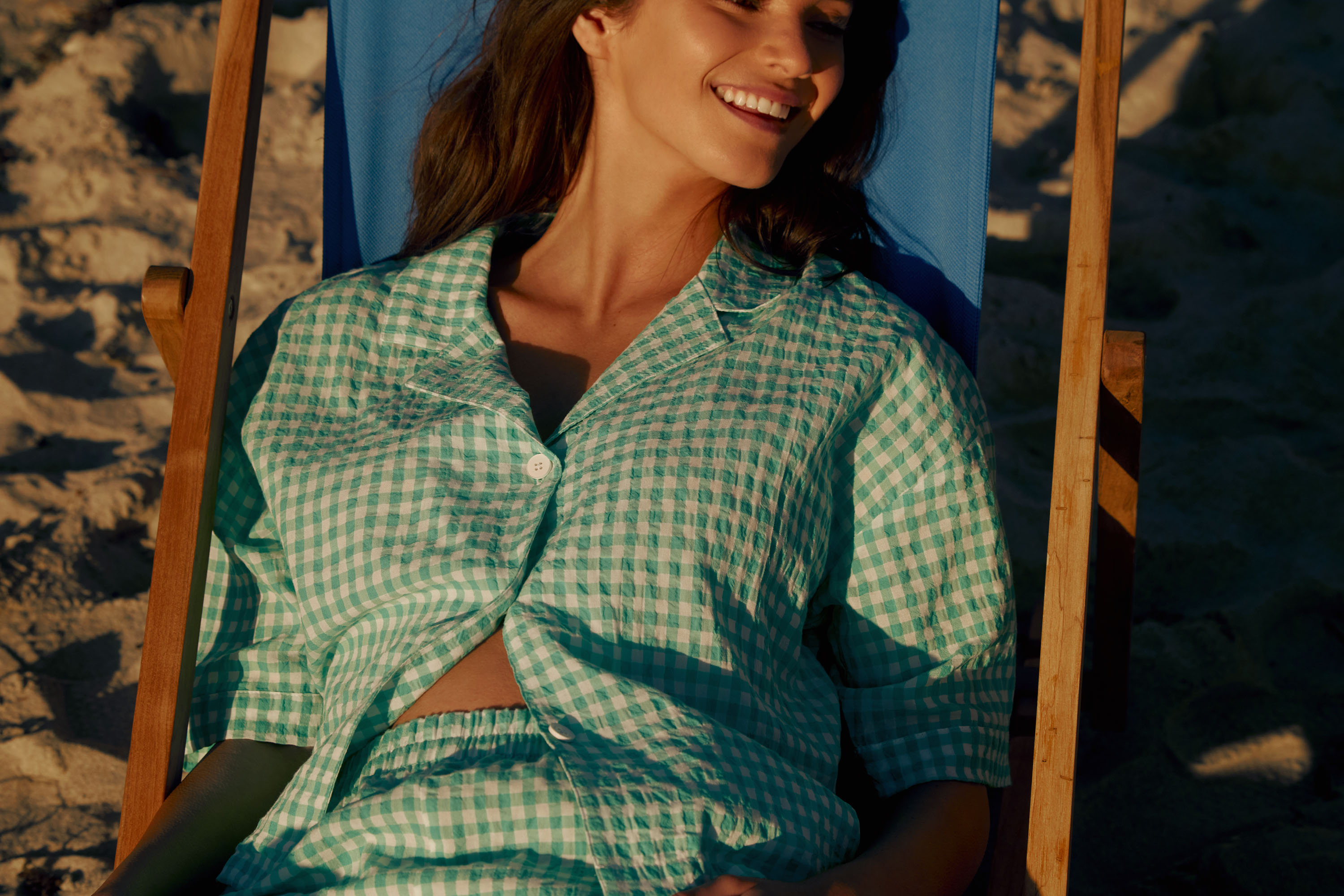 Upper body image of a young lady wearing gingham green pyjamas, lounging on a beach chair. 
