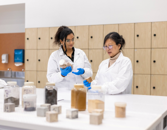 Two females in a lab environment working together. They are wearing white coasts and blue gloves.