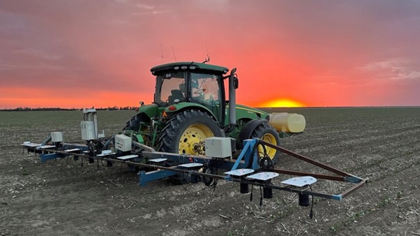 A green John Deere tractor fitted with a mounted spray boom and yellow tank, parked in a ploughed field at sunset with an orange sky behind.
