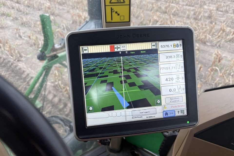 A guidance screen mounted in a tractor cab, showing a green field map with coverage data, with a paddock and green machinery visible through the window.