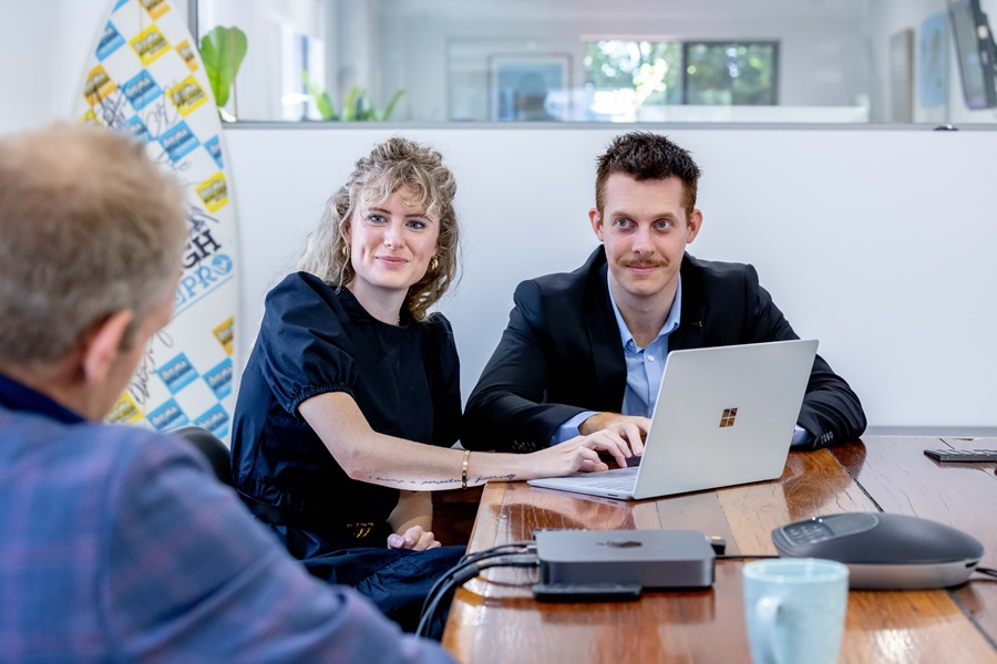 A smiling man and woman facing the camera talk with an suited man over laptops in a meeting room