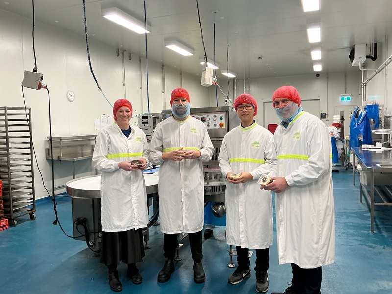 A group of four people wearing white lab coats and red hair nets inside a food manufacturing plant