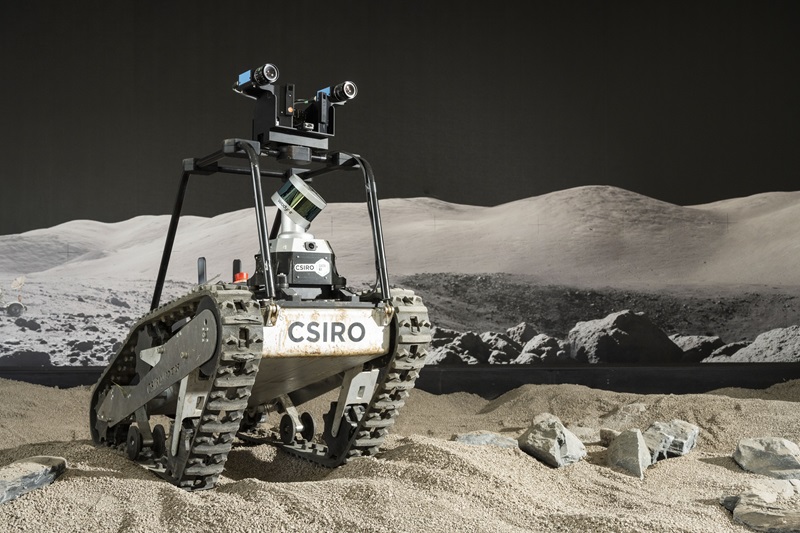 a bi-focal camera sits on top of a tracked robot in a sandpit-like space that is filled with crusher dust to simulate the lunar landscape. An image of the real lunar landscape is on the wall in the background.