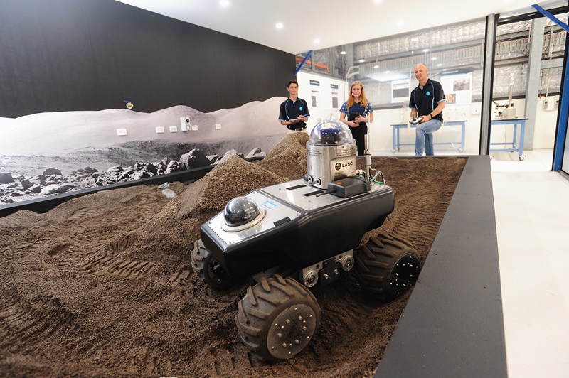 A four-wheeled rover sits in the foreground in a sandpit area filled with crusher dust to mimic the Moon's surface. Three people stand in the background controlling the rover. They are all inside a room which has a Moon scape image on the far wall. 