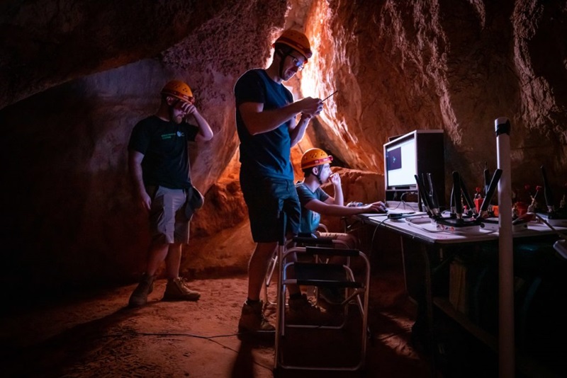 Three male human operators wearing hardhats in a cave control a robot and interact with a computer interface