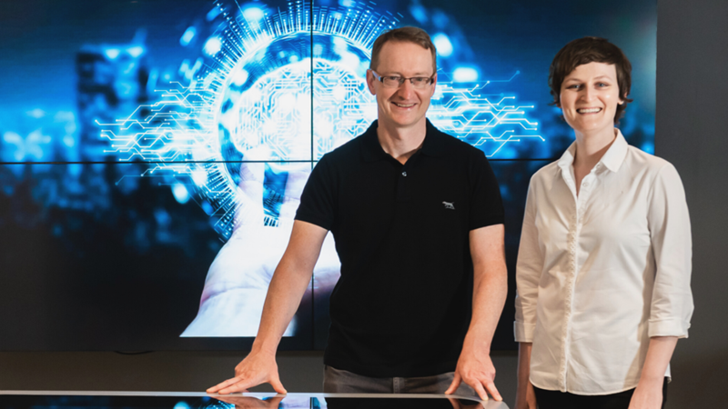 Man in black shirt wearing glasses and woman in white shirt stand in front of blue screen.