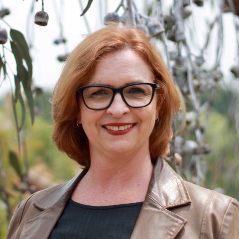A professional photo of Dr Melanie McGrath. She is standing in front of a gum tree with leaves in the background. She wears black rimmed glasses, a brown leather jacket and is smiling at the camera