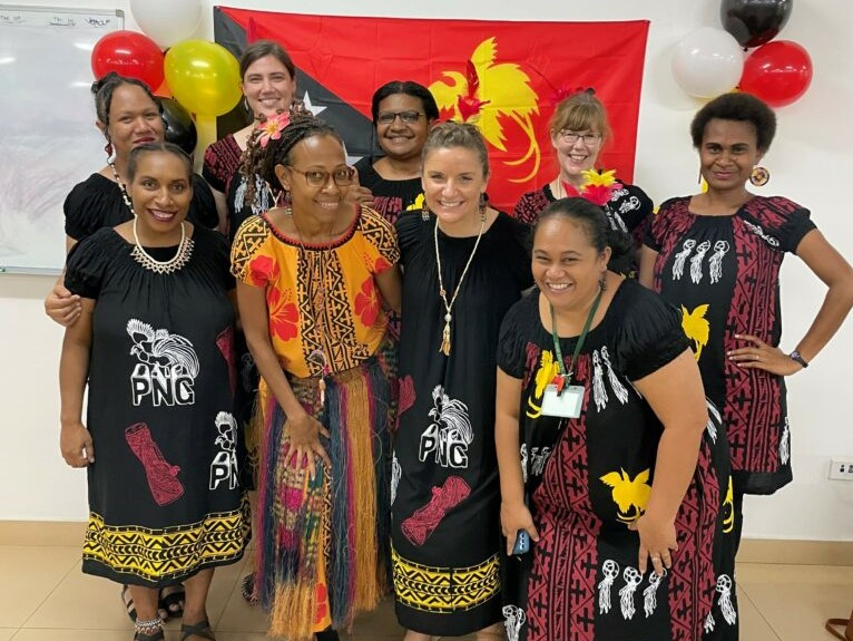 A group of people all looking at the camera and smiling. In the background is the flag of Papua New Guinea and balloons.