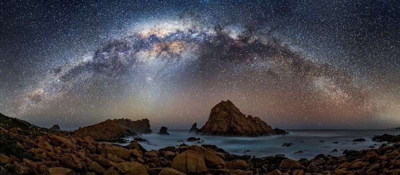 A astrophotograph of stars in the nights sky with an ocean in the foreground at night time.