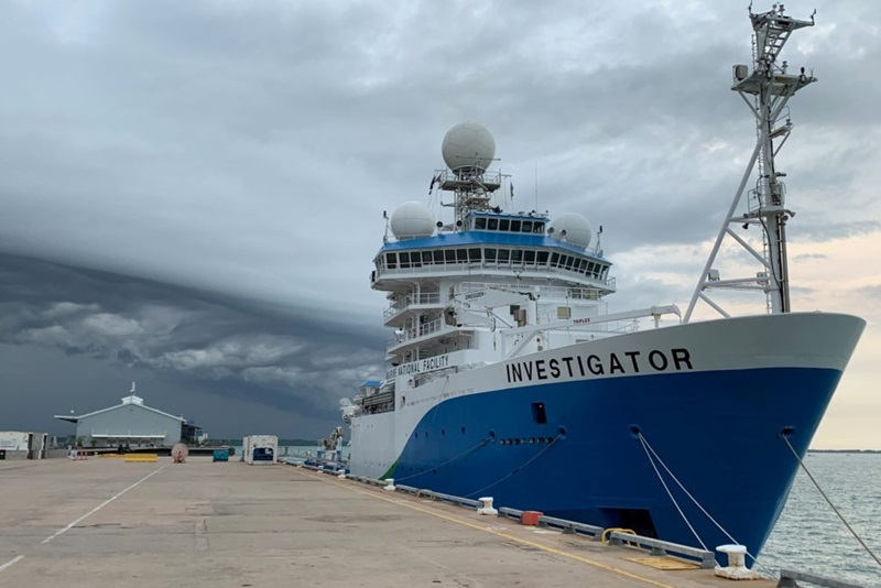 A large blue and white ship at a wharf with a storm forming in the background.