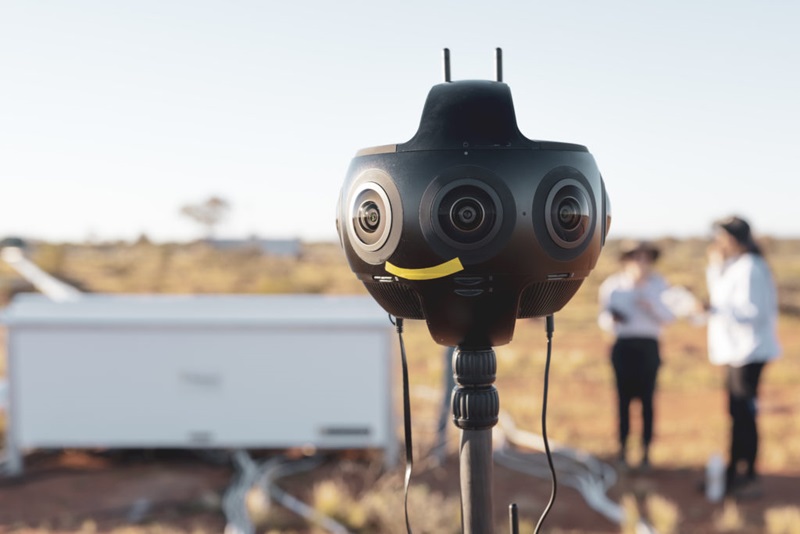 A circular camera with many lenses is in the foreground centre of the image in sharp focus, while the background is out of focus and vaguely shows two women talking to the right of the camera and a red dirt landscape with a pale blue sky.
