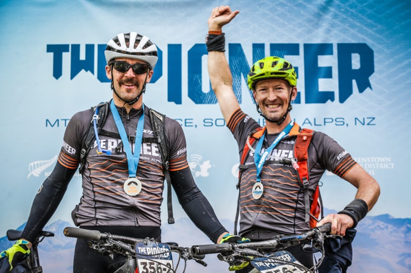 Two men in riding gear. One man has his right arm in the air.