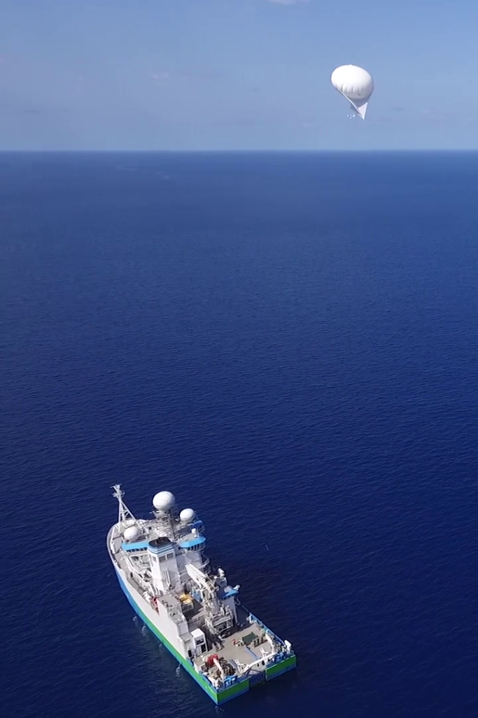 An aerial view of a ship on the ocean with a white balloon floating above it.