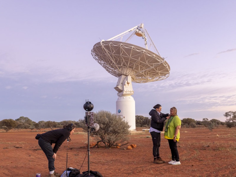 A landscape of red dirt and a pale blue sky with a large white radio telescope dish in the centre-background. The foreground has a young woman bending over a piece of filming equipment and an unusual many-eyed camera on a tripod to the left and another pair of women to the right with one adjusting the microphone attached to the scarf of the second woman.