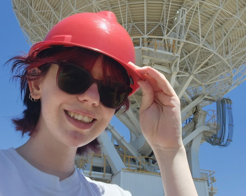 A young woman wearing sunglasses and a red hard hat, smiling underneath a large satellite dish.