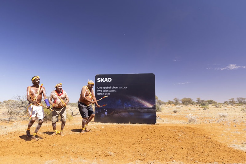 Three Indigenous men (Godfrey Simpson, Geoffrey Mongoo and Gerard Boddington) who are painted up are performing a Wajarri cultural dance in front of an SKAO sign. The sign reads: "SKAO One global observatory, two telescopes, three sites"