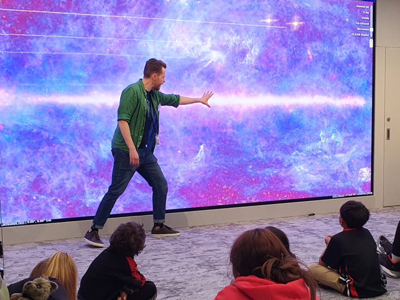 A man standing an motioning his hand at a screen behind him while kids watch. The screen shows a flurry of purple, blue and pink colours with a lighter horizontal line in the middle. 