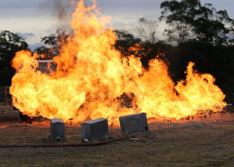 Burnovers testing on a firetruck. A bush scene with large flames. there are three metal boxes in the foreground