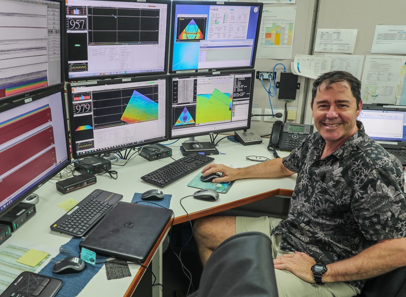 A man sitting at a desk with six computer screens showing map data