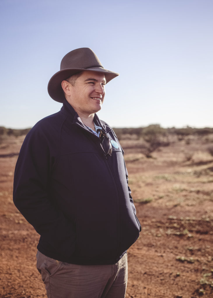 A man in a hat and jacket to the left of the frame against a background of red dirt and pale blue sky. 