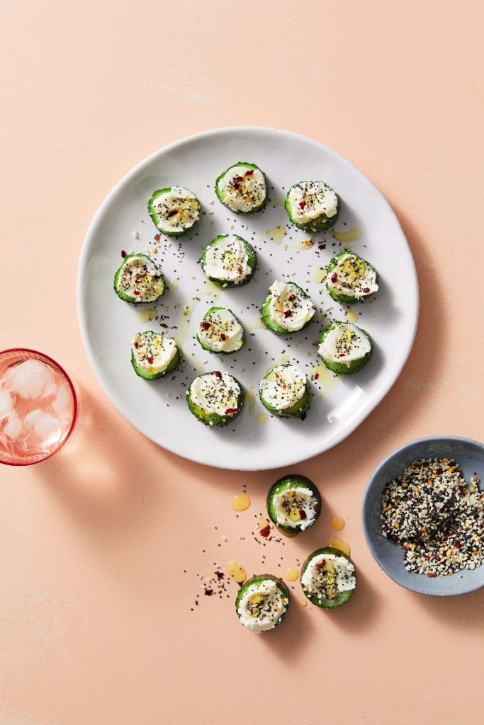 On the salmon pink kitchen top bench cucumber slices, with cream cheese and seeds and spices sit on a plate with olive oil drizzled over them. Next to the plate is the seed and spice mix in a grey bowl. On the kitchen top bench between the two some of the Cucumber Cream Cheese Bites are laid out too