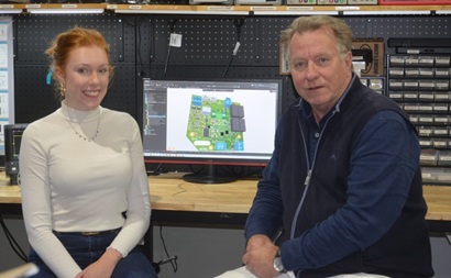 Jasmine and John sitting at a workbench smiling at the camera. There is a computer screen showing an animated circuit board in the background. 