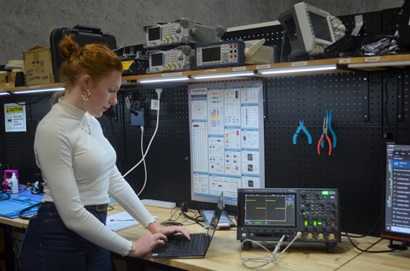 Jasmin standing at a work bench typing on a laptop. Her laptop is connected to an engineering machine on the bench.