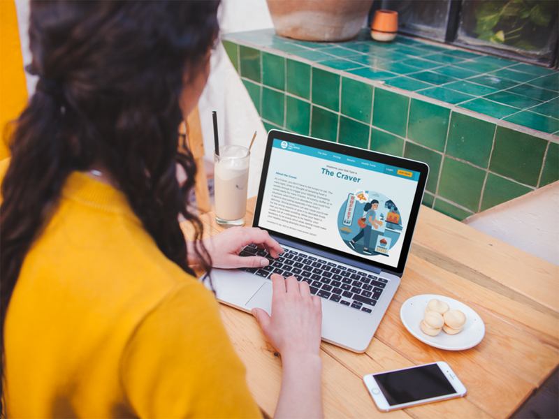 A person is sitting at a bench with a laptop open. She is reading off the screen. Next to her on the bench there is a small plate of biscuits, a mobile phone and a glass of ice coffee.