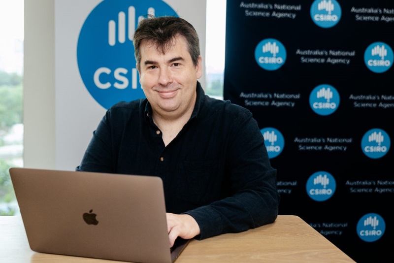 A middle-aged man sitting at a laptop in front of a CSIRO-themed media wall, smiling.