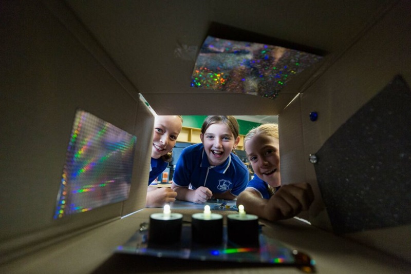 Three primary-school age kids looking excitedly into a cardboard box with three small lights at the base