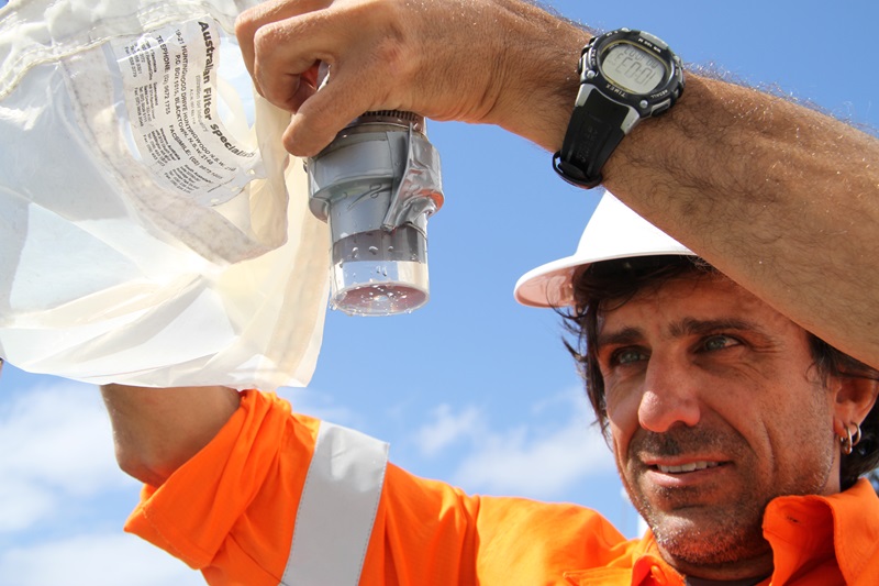 Educator on Board Chris in high-visibility orange shirt and hard hat looking at a water sample in a container