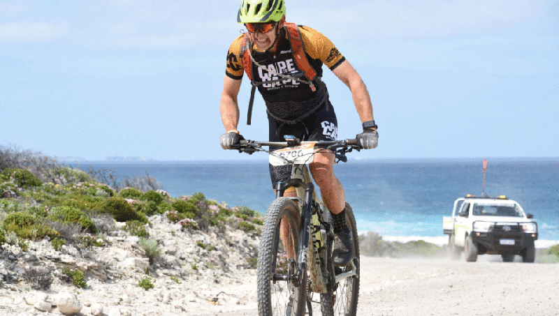A man riding a bike on a dirt road with a 4WD and a beach behind him