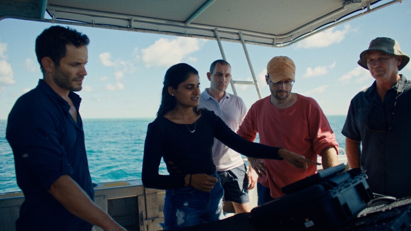 Four men and one woman stand on a boat on blue water discussing AI in conservation. The woman is pointing at a screen out of sight.