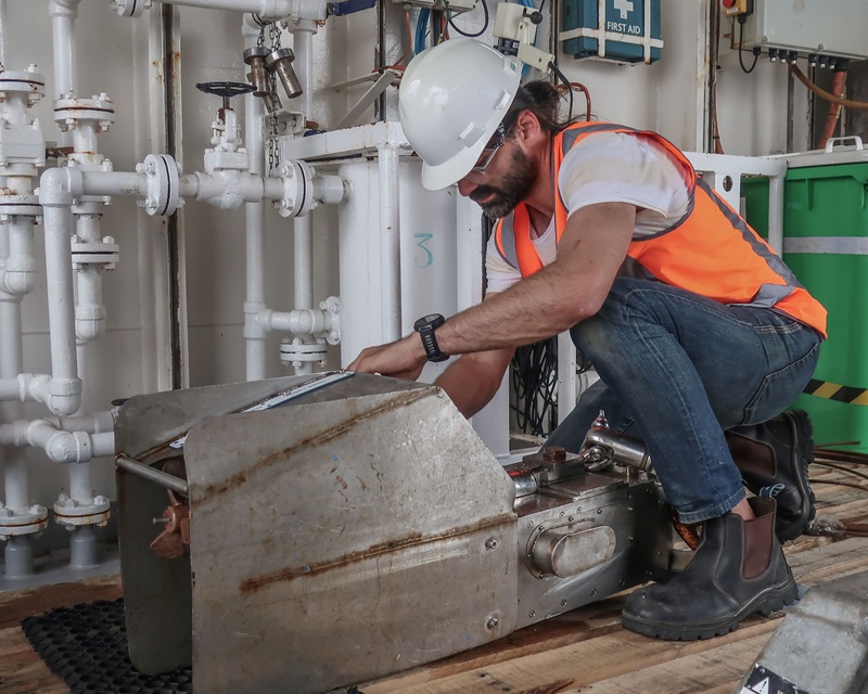 A man in high-visibility vest and hard hat leaning over a metal box. there are pipes and valves in the background