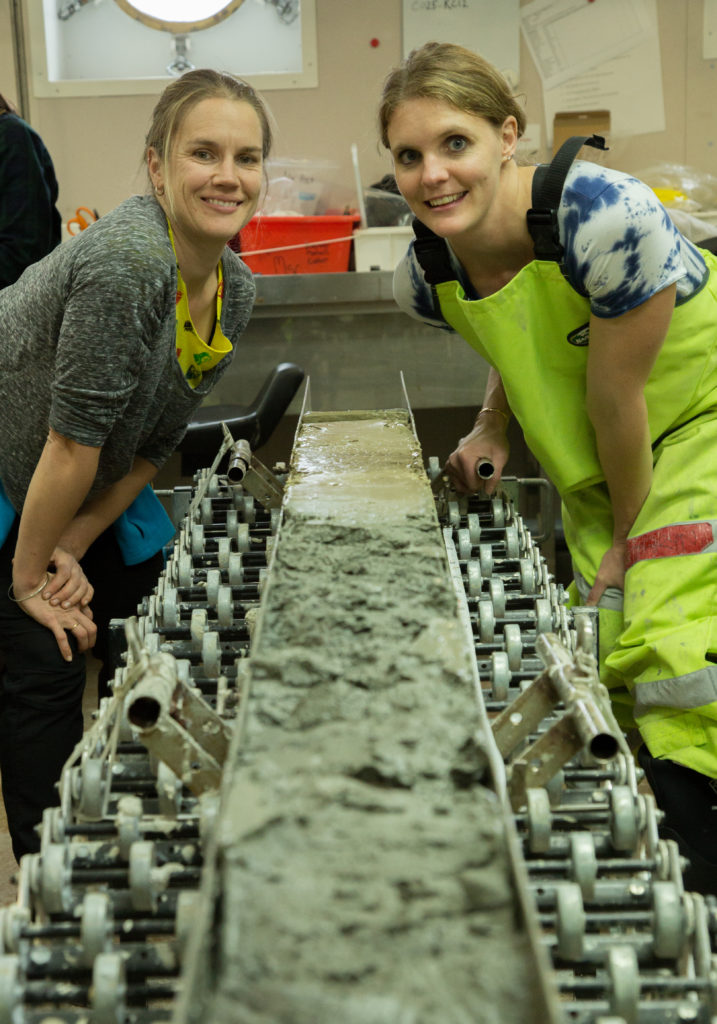 Two people standing either side of a tube filled with mud smile at the camera.