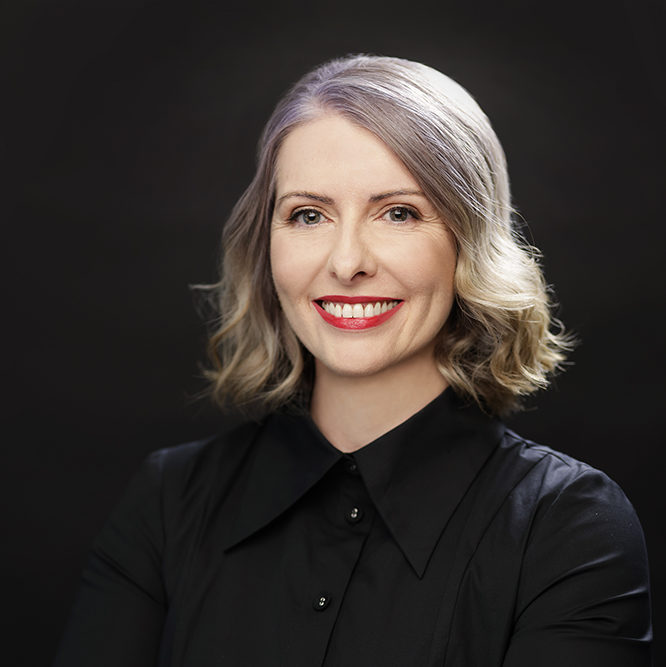 Justine Lacey smiling at the camera wearing black shirt and red lipstick. She is standing in front of a black background. 