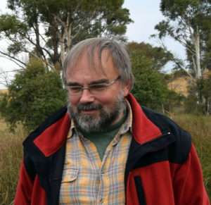 A middle aged man wearing a red jacket, standing outside surrounded by gum trees.