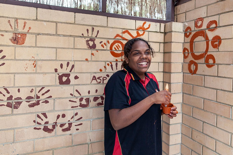 A young female student is smiling at the camera and is holding a small cup filled with paint and a paintbrush. Behind her is a brick wall upon which paintings have been made by students including handprints.
