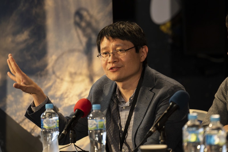 A man (Mark Cheung) can be seen sitting behind a table which has a microphone on it. He is speaking to an audience who is behind the camera.