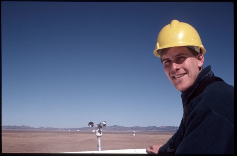 A man in a yellow hard hat stands on top of a large antenna in the desert.