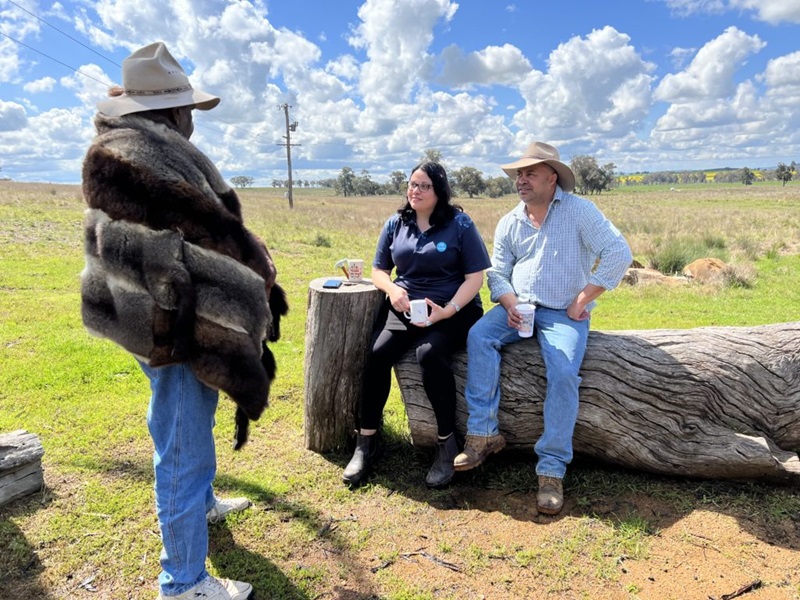 One person standing talking to two people sitting on part of a large fallen tree in a paddock,
